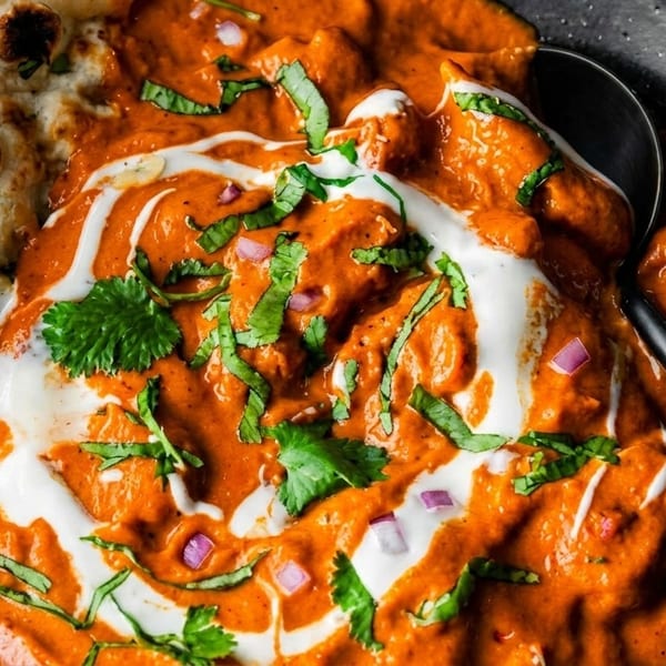Close-up of butter chicken with naan bread, garnished with cream, cilantro, and red onions.