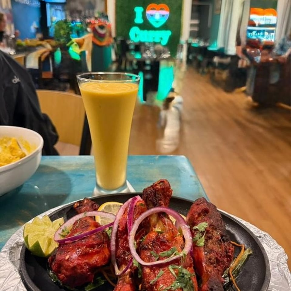 Indian restaurant table with assorted grilled kebabs, red onions, lime, cilantro, and a tall mango lassi beneath an “I Love Curry” sign.