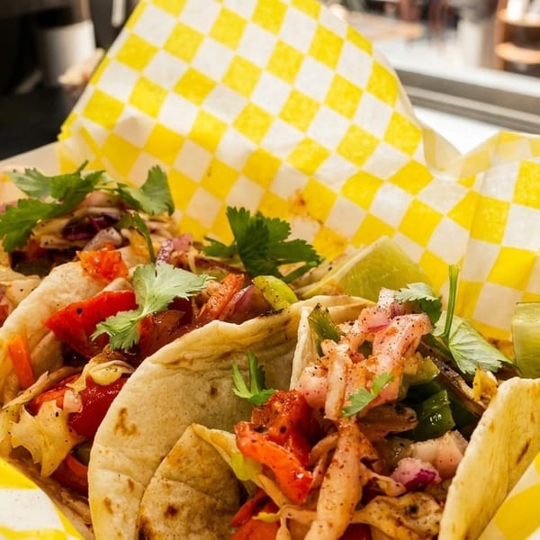 Three vibrant tacos with various fillings and cilantro, served in yellow checkered paper.