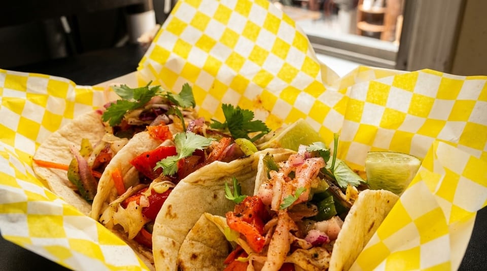 Three vibrant tacos with various fillings and cilantro, served in yellow checkered paper.