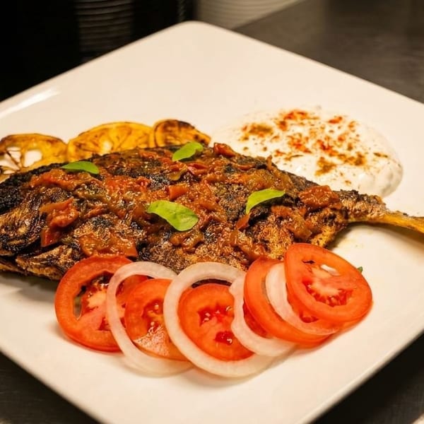 Whole grilled fish topped with tomato sauce, served with lemon, yogurt, and sliced tomatoes and onions.
