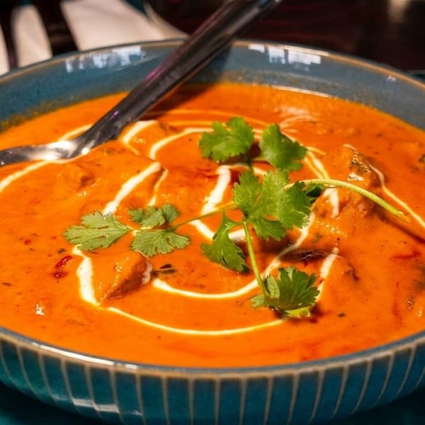 A blue bowl of butter chicken curry with cream swirls, cilantro, and a spoon.
