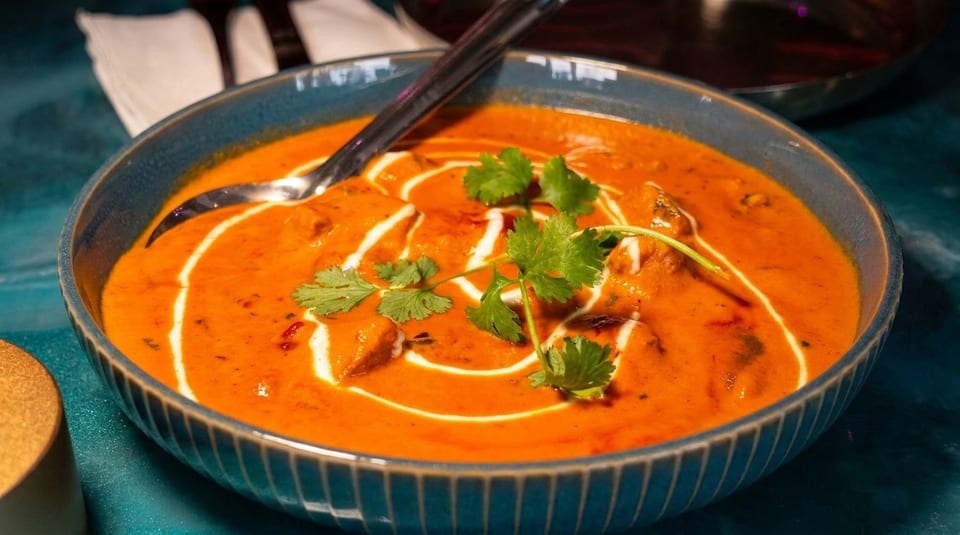 A blue bowl of butter chicken curry with cream swirls, cilantro, and a spoon.