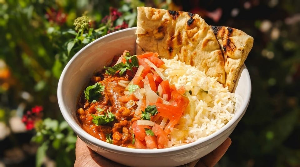 A bowl of curry with rice, tomato and onion relish, and naan bread, held outdoors.