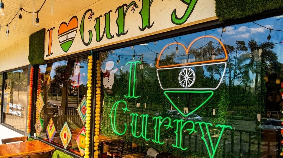 The exterior of I Curry restaurant with a neon sign and Indian flag heart in the window.
